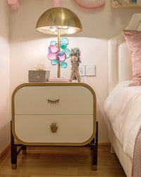 Lit mushroom lamp with pink and teal acrylic orbs on a cream nightstand beside a bed in a soft pink room.