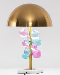 Front view of the colorful acrylic table lamp with a brass dome, pastel pink, blue and green spheres, marble base and visible power cord.