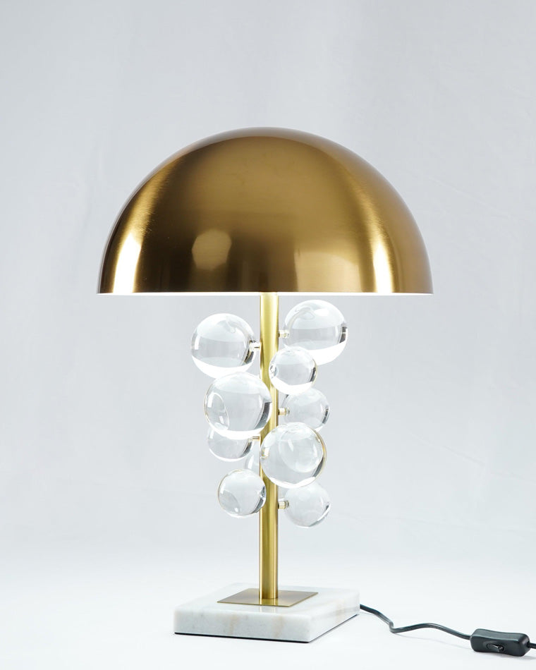 Studio view of the clear acrylic table lamp with transparent spheres, brass dome and stem, white marble base, and inline cord switch.