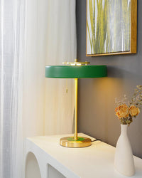 Green mid century modern table lamp glowing on a white console beside sheer curtains and a tall vase of dried flowers.