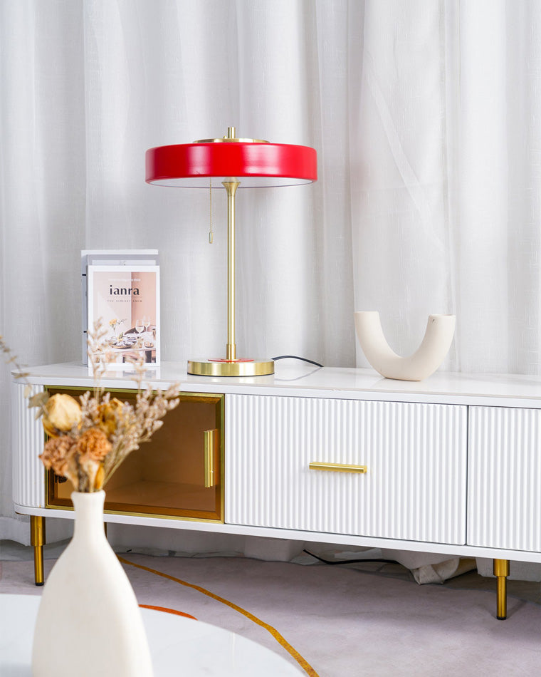 Red metal table lamp with a wide circular shade and gold-tone frame on a white console against sheer curtains.
