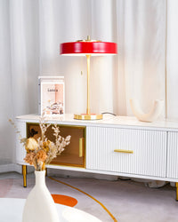 Red modern table lamp glowing on a white fluted console, framed by dried flowers and sheer curtains.