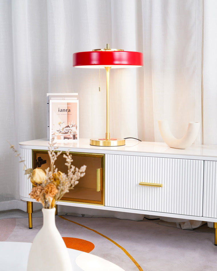 Red modern table lamp glowing on a white fluted console, framed by dried flowers and sheer curtains.