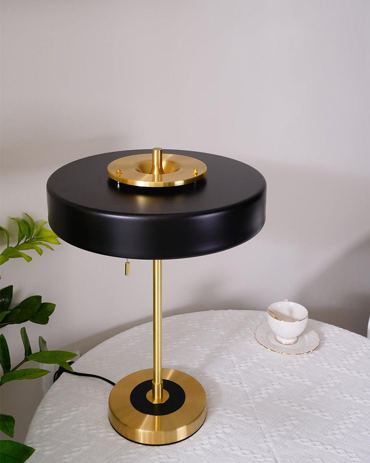 Unlit black metal table lamp with a broad circular shade on a covered table beside a teacup and plant.