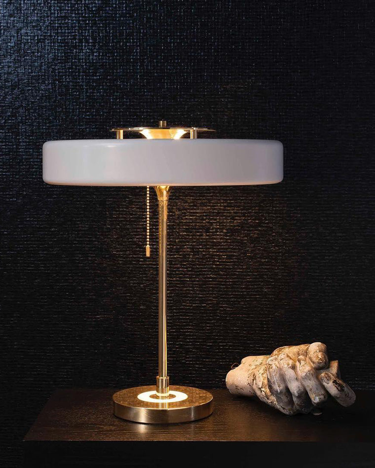White led table lamp illuminated against a textured black wall, showing its broad disc shade and gold-tone pull-chain frame.