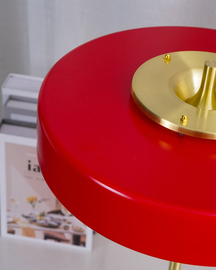 Close-up of a red metal table lamp showing the smooth round disc shade and circular gold-tone hardware on top.