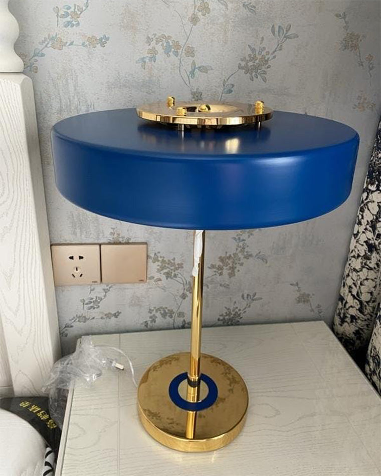 Blue mid century modern lamp with a round shade and gold-tone frame on a white bedside surface against floral wallpaper.