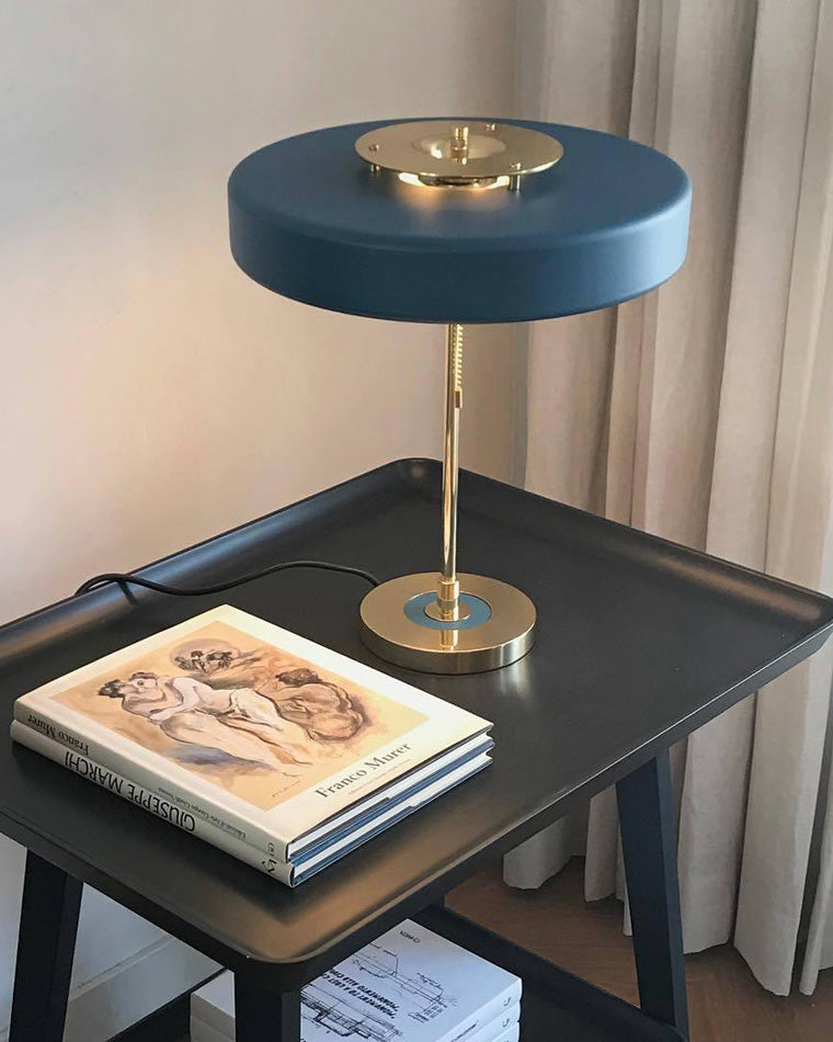 Blue desk lamp with a wide round shade and gold-tone base on a black tray table beside stacked art books.