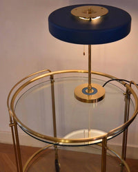 Blue pull chain table lamp glowing on a round glass and gold-tone trolley, with the hanging chain and power cord visible.