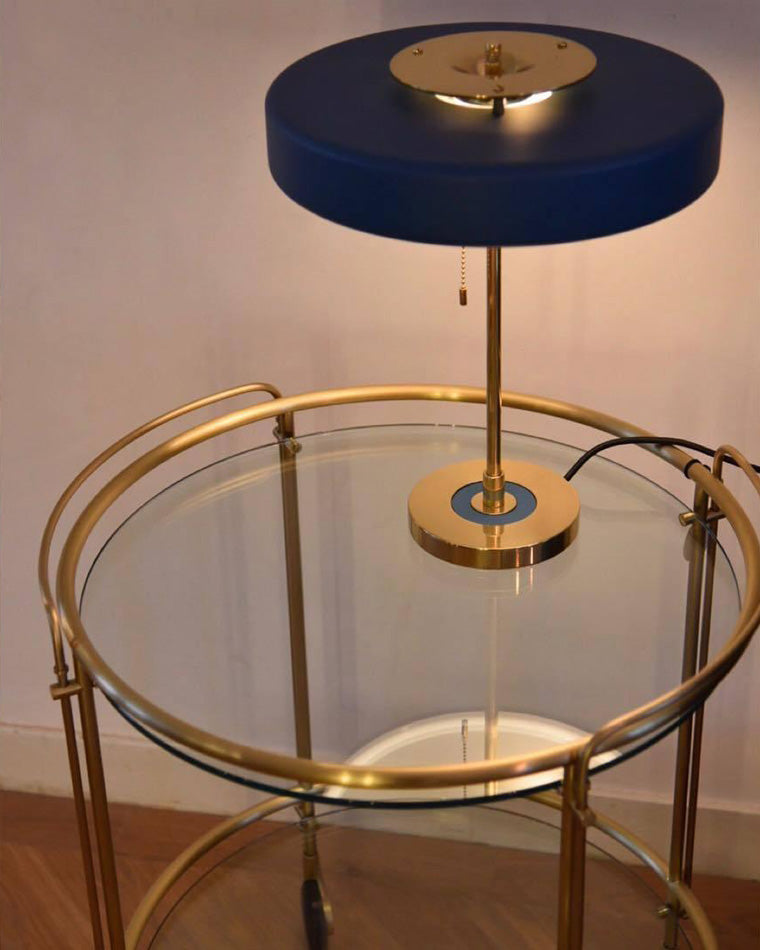 Blue pull chain table lamp glowing on a round glass and gold-tone trolley, with the hanging chain and power cord visible.