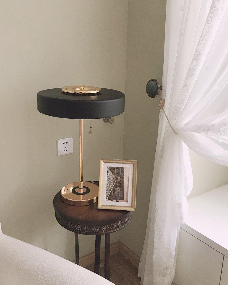 Black vintage table lamp on a carved dark-wood side table beside a framed print and sheer window curtain.