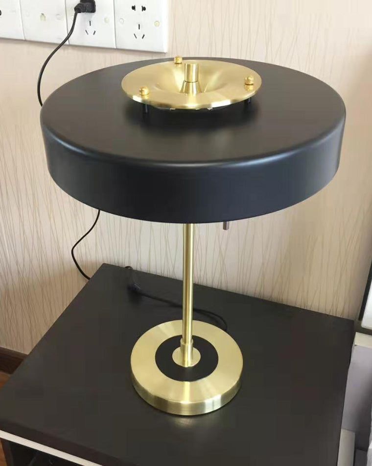 Black table light on a dark nightstand, showing its broad round shade, gold-tone base and plugged-in power cord.