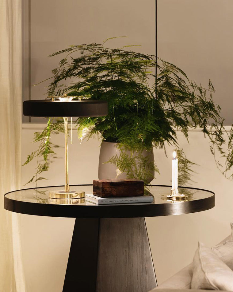 Black mid century table lamp glowing on a glossy round table with a large fern, stacked books and a lit candle.