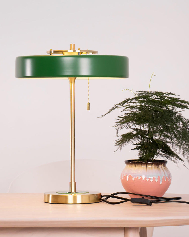 Green pull chain table lamp beside a potted plant, showing the round gold-tone base, hanging chain, cord and inline switch.