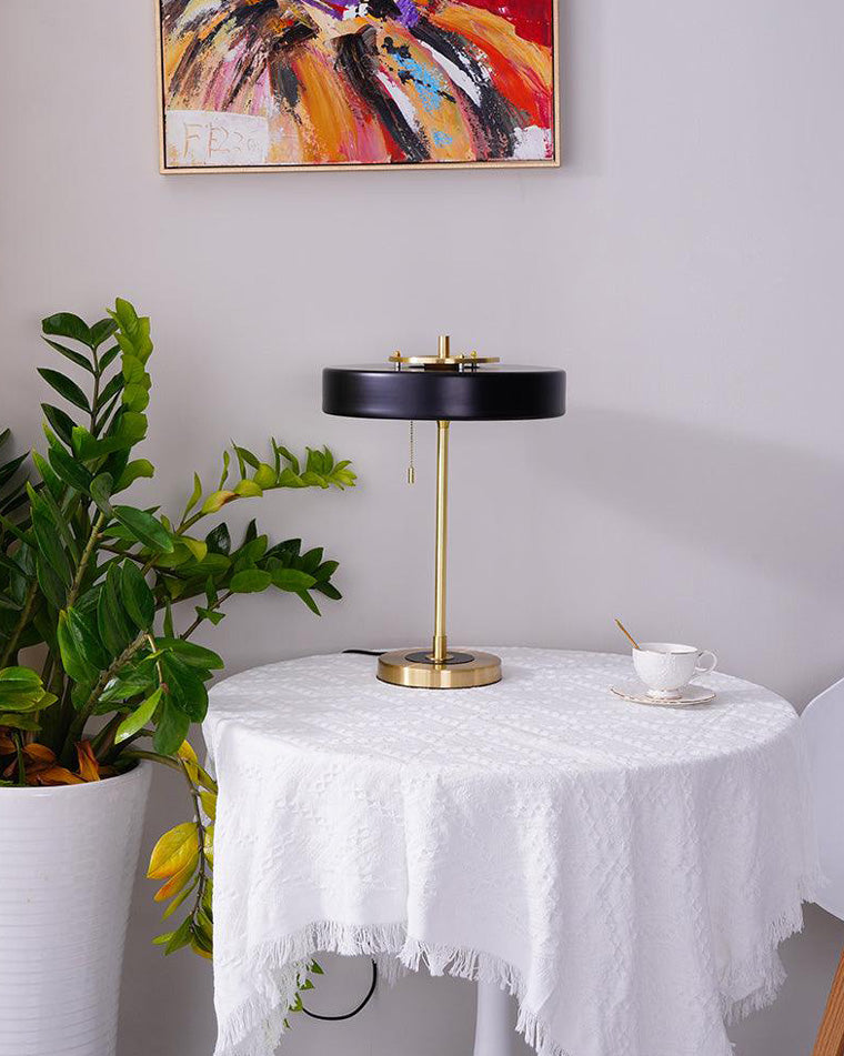 Black vintage desk lamp on a round covered table beside a leafy plant, teacup and colorful abstract artwork.