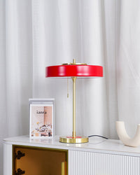 Red table light with a wide metal shade and gold-tone frame on a white fluted console beside books and a curved ceramic object.
