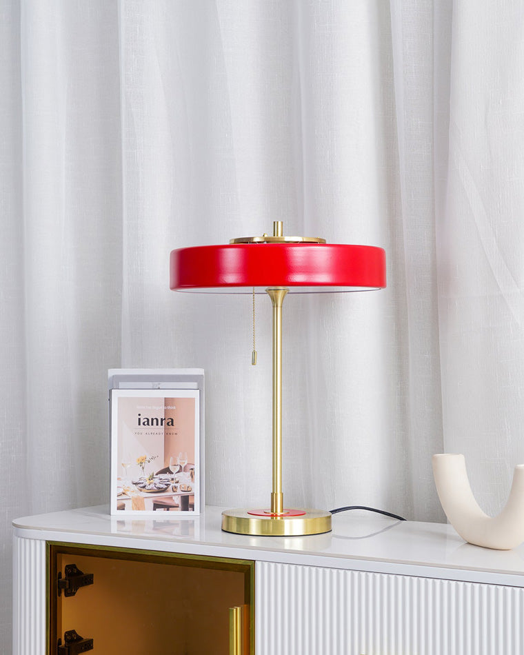 Red table light with a wide metal shade and gold-tone frame on a white fluted console beside books and a curved ceramic object.