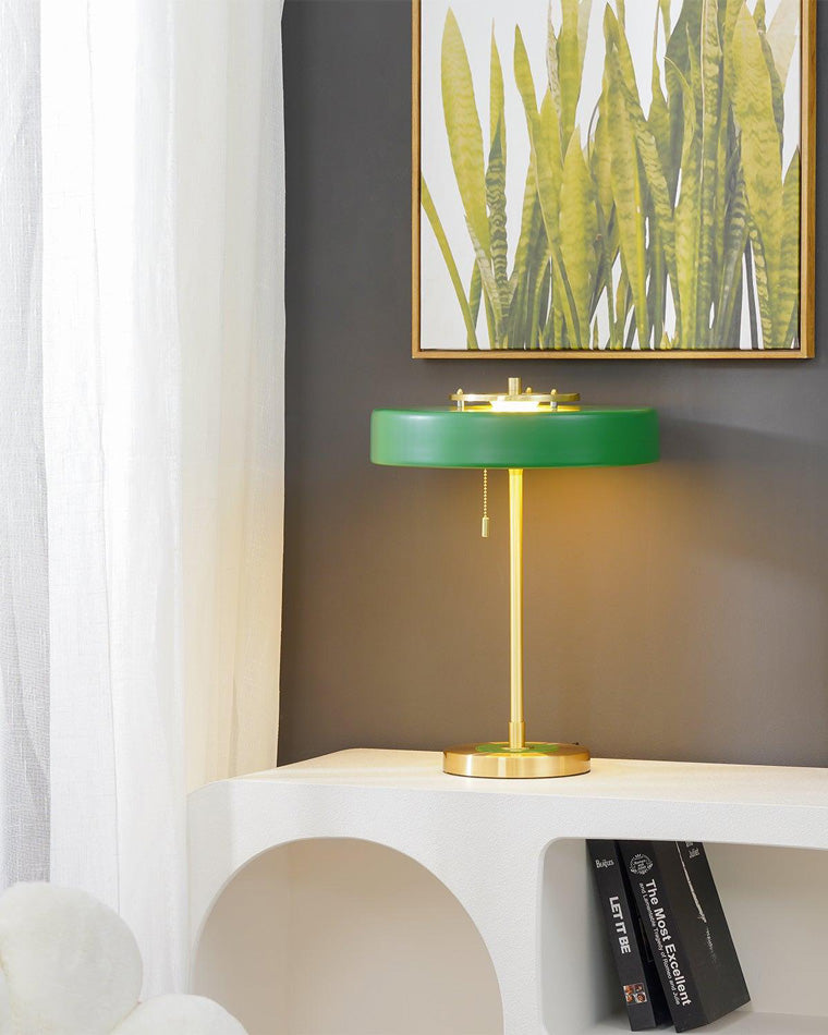 Green mid century table lamp illuminated on a white console beneath botanical artwork, casting warm light on a dark wall.