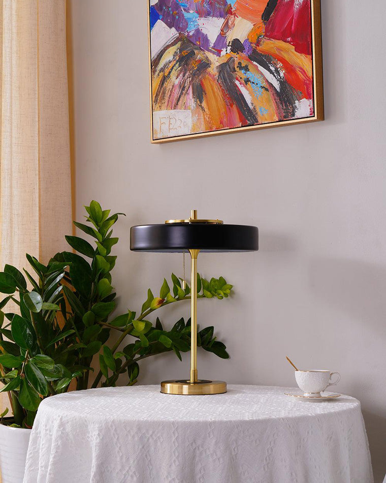 Black vintage table lamp on a round covered table beside a large plant, teacup and colorful abstract painting.