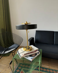 Black mid century modern lamp on a green wire side table with stacked books, framed by black seating and olive curtains.