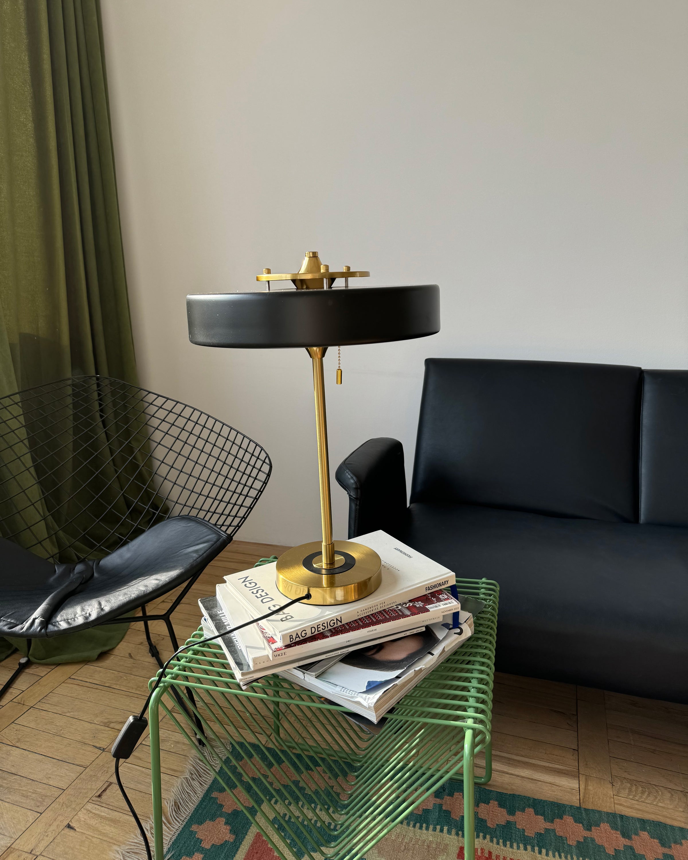Black mid century modern lamp on a green wire side table with stacked books, framed by black seating and olive curtains.