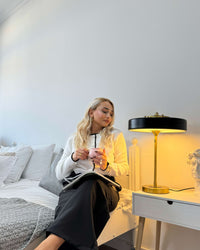 Black table light with a gold-tone frame glowing on a white nightstand beside a seated woman holding a cup.