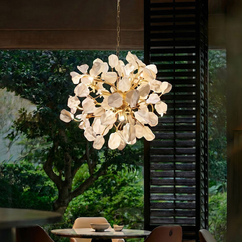 Square view of a dining table chandelier with white ceramic petals and brass branches glowing above a round table beside lush garden windows.