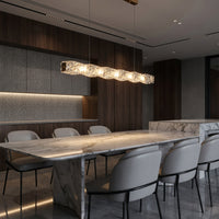 Lit linear chandelier dining room fixture with a twisted crystal bar above a long marble table in a dark wood interior.