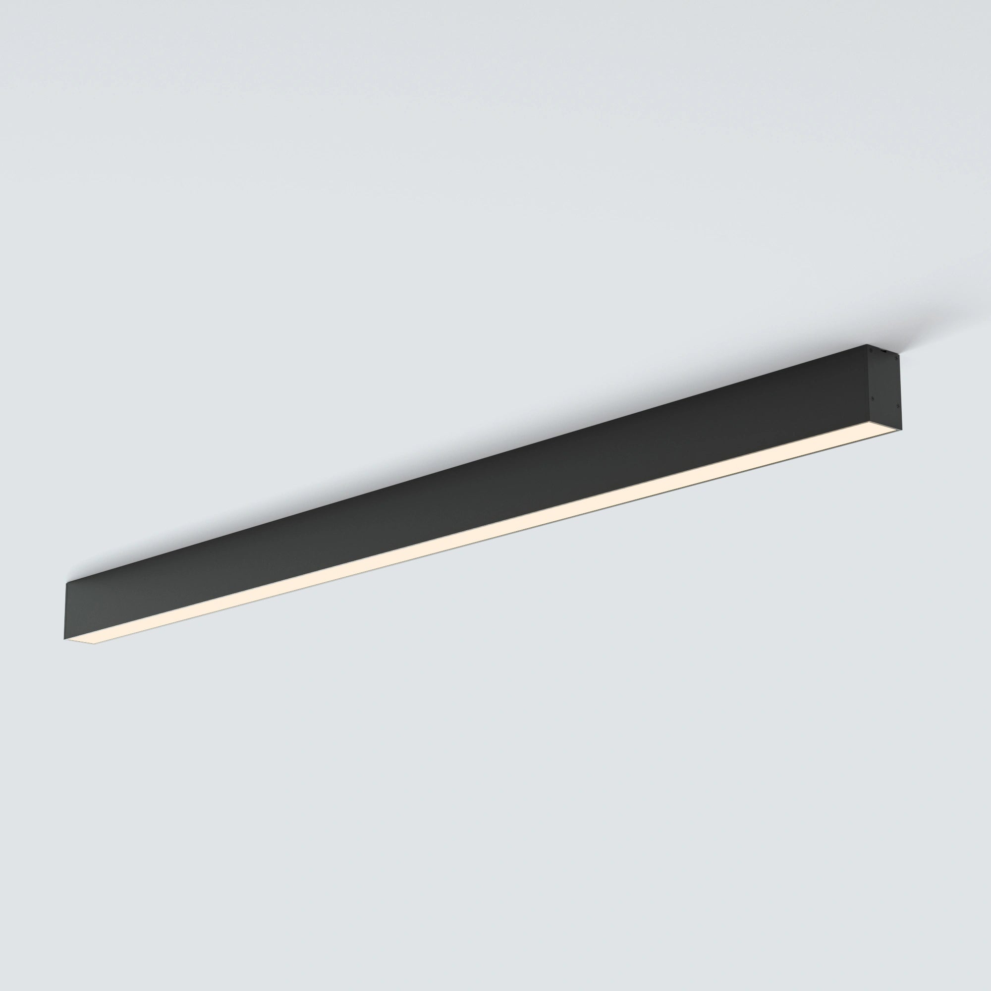 A modern linear LED ceiling light fixture in matte black, shown mounted against a plain light gray wall with the light turned on, emitting a warm white glow.