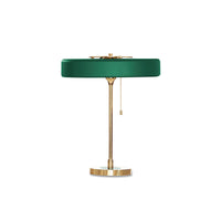 Green metal table lamp with a flat round shade, gold-tone frame, circular base and hanging pull chain on white.