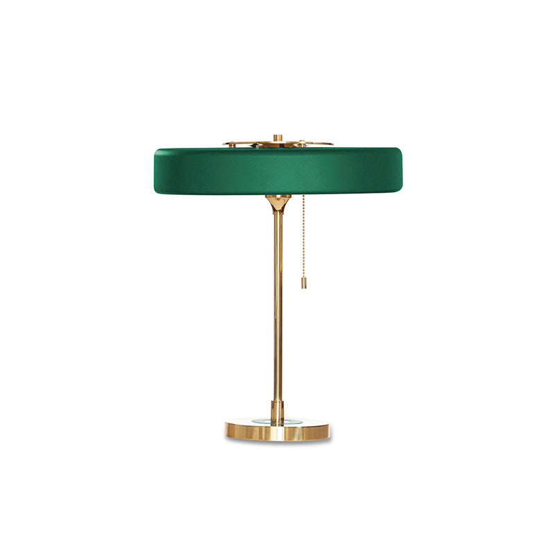 Green metal table lamp with a flat round shade, gold-tone frame, circular base and hanging pull chain on white.