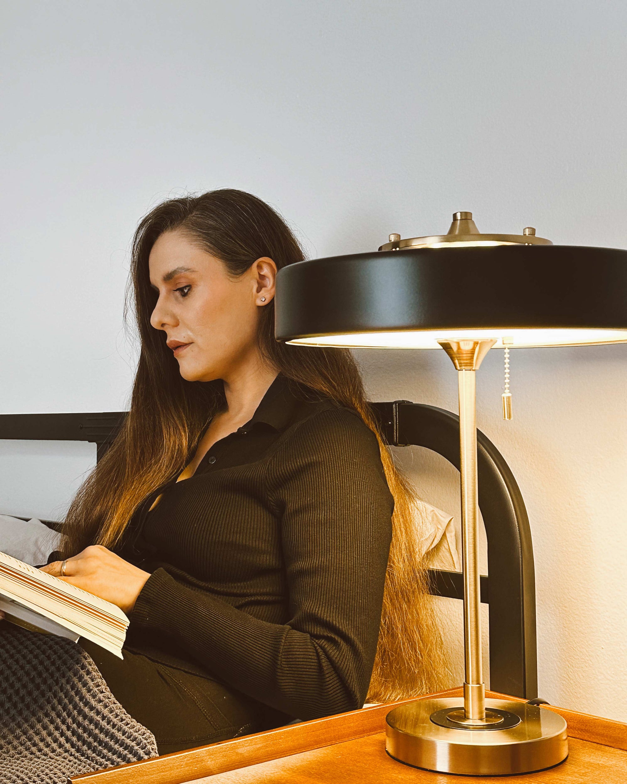 Black pull chain lamp casting warm downward light beside a woman reading in bed.