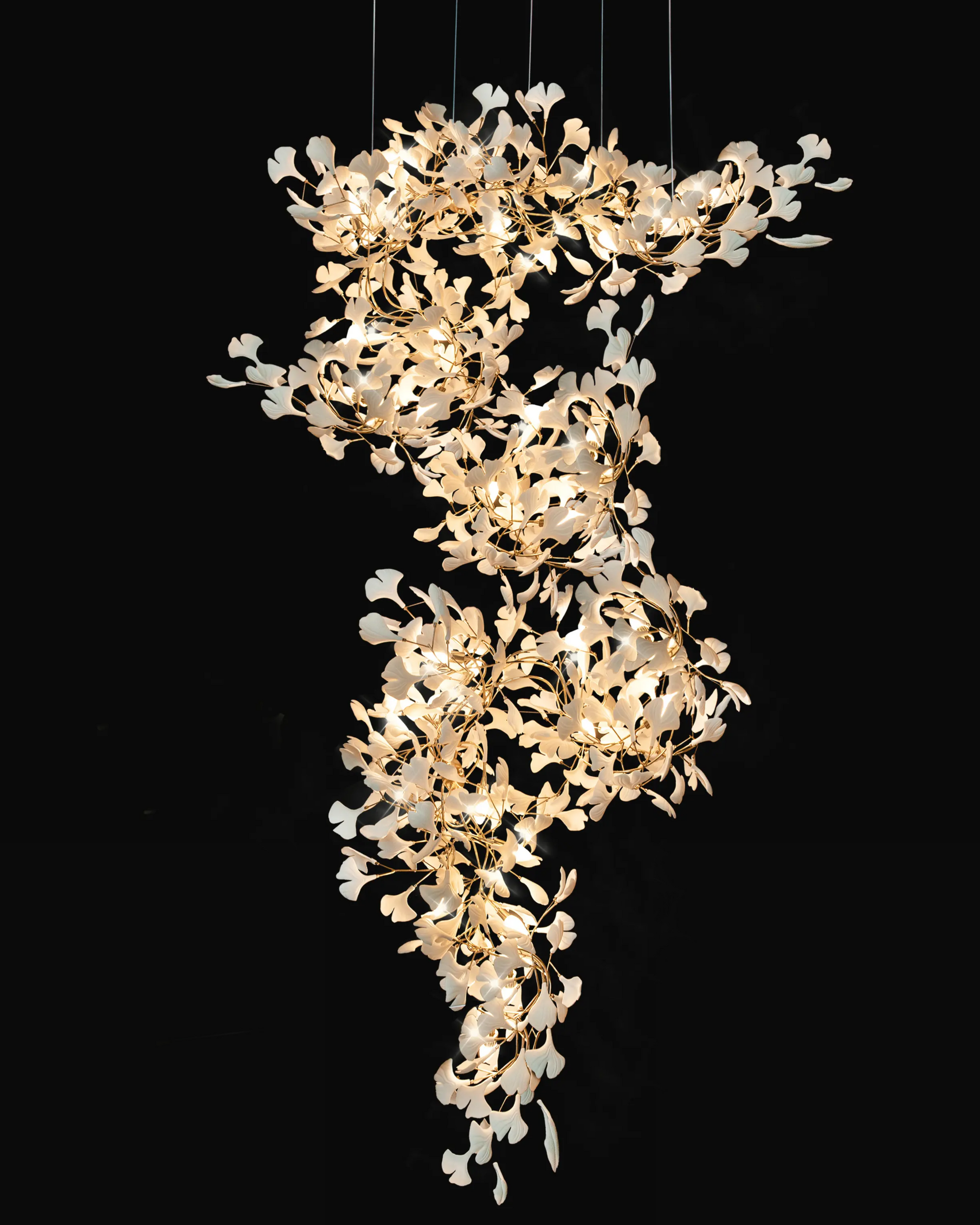 Illuminated ceramic ginkgo branch chandelier with glossy white leaves and gold-toned brass branches against a black background.