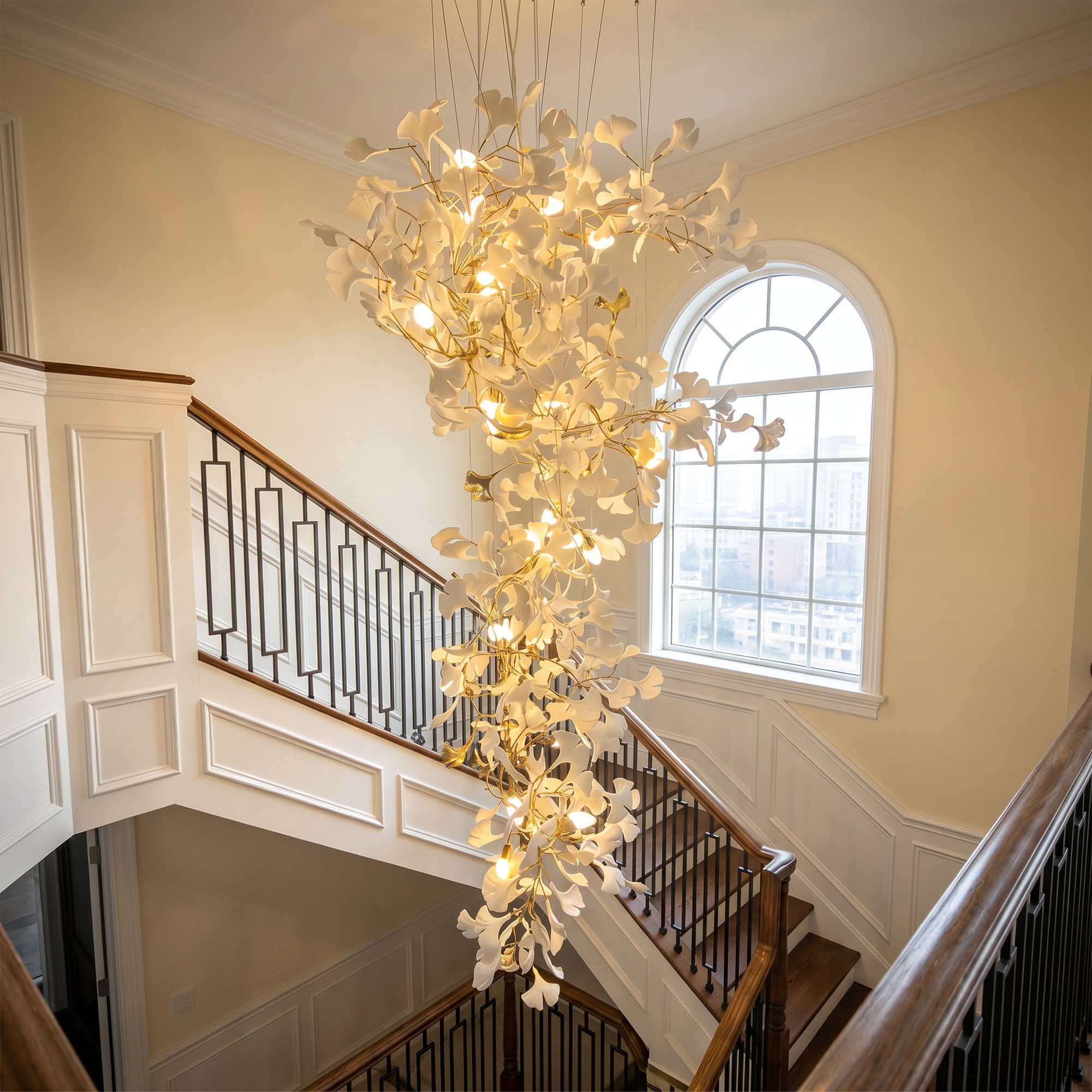 Modern staircase chandelier with illuminated ceramic ginkgo leaves hanging beside a tall arched window.