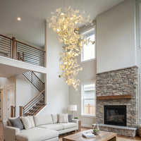 High ceiling chandelier with white ceramic ginkgo leaves glowing above a double-height living room with a staircase and stone fireplace.