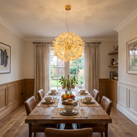 A modern spherical white and gold chandelier with multiple ceramic ginkgo leaf pendants hangs above a wooden dining table set for six in an elegant dining room.